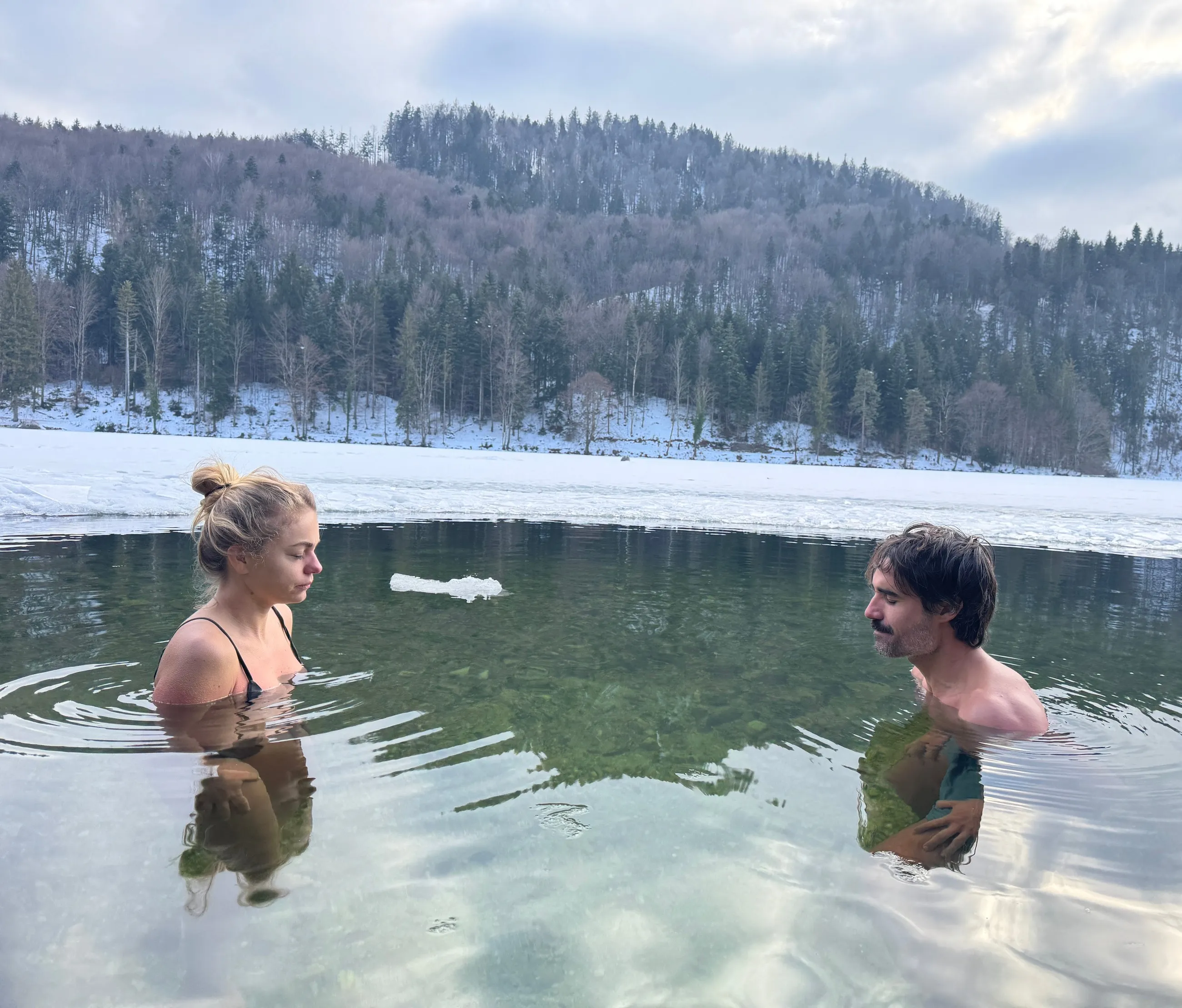 Zwei Teilnehmende sitzen bis zur Brust in einem See, auf dem Wasser treiben Eisschollen, dahinter verschneiter Wald
