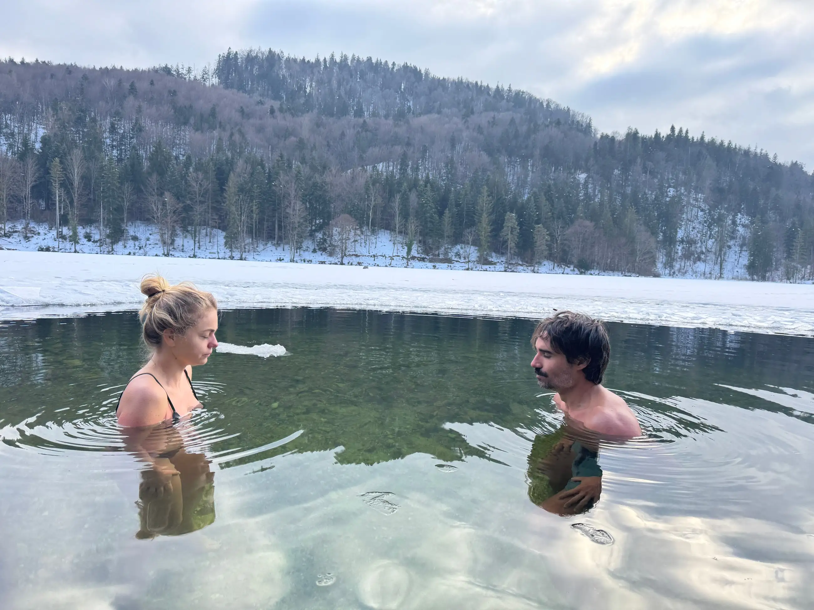 Zwei Menschen stehen sich in einem winterlichen Bergsee gegenüber, dahinter verschneiter Wald