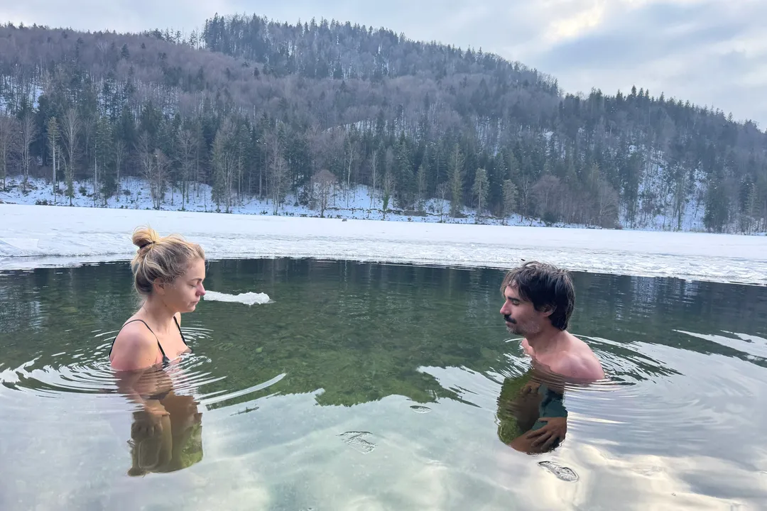 Zwei Teilnehmende sitzen bis zu den Schultern in einem eisigen Bergsee, dahinter Schnee und Wald
