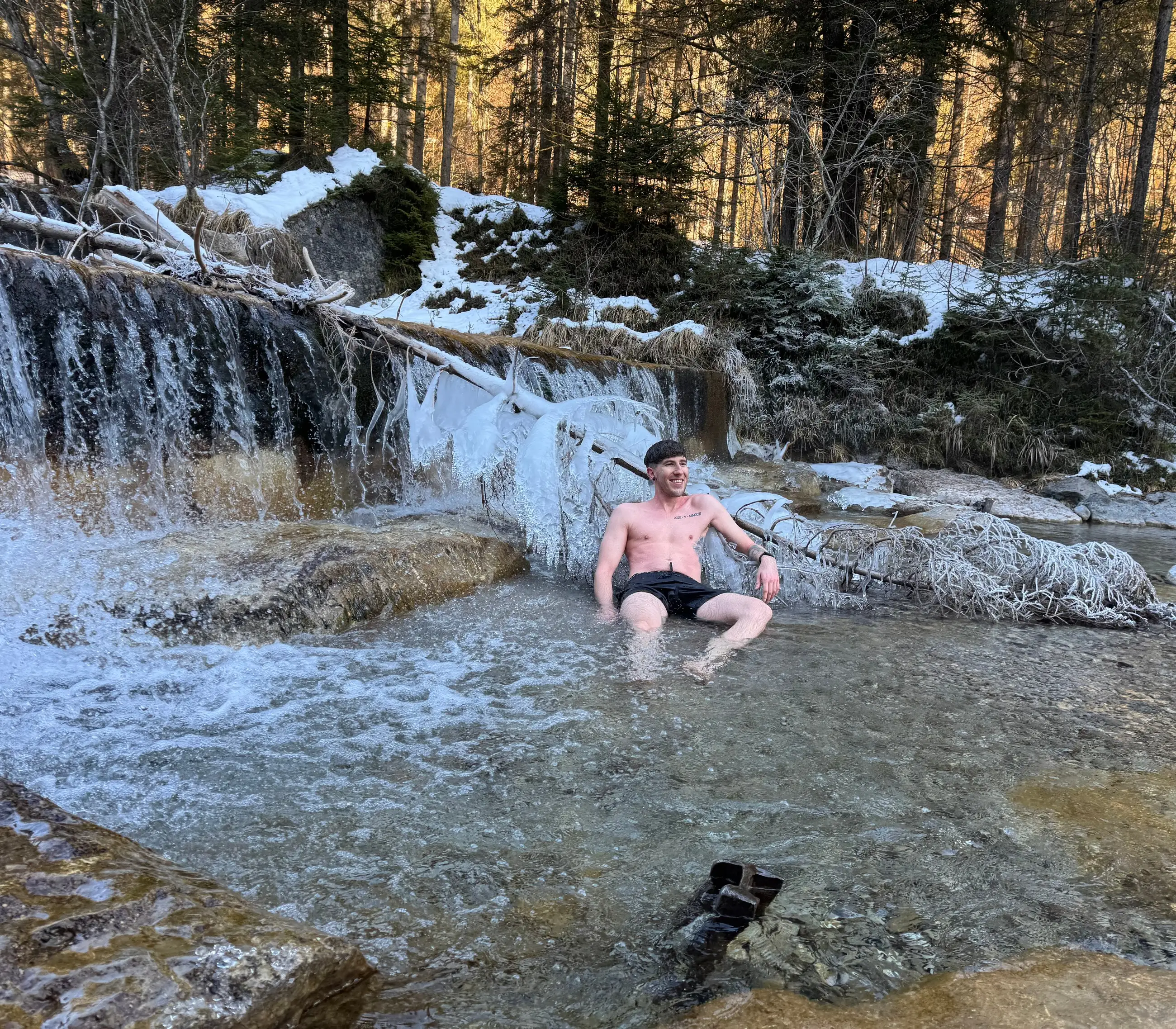 Julian Scheuermann sitzt im Winter in einem eiskalten Bach unterhalb eines vereisten Wasserfalls